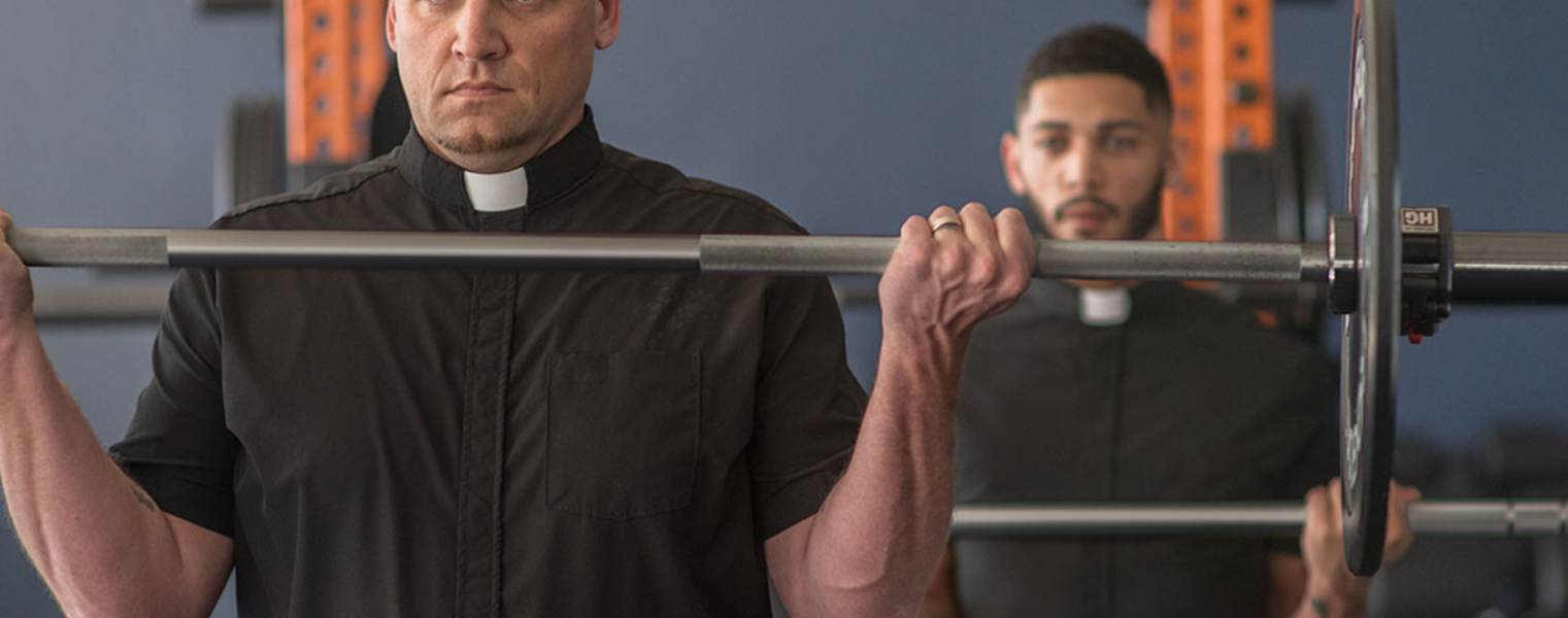 two men wearing the performance clerical shirts from wicking vicar both curling barbells in the gym