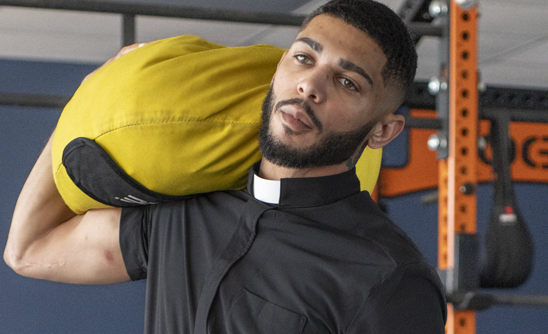 man wearing a short sleeve clergy shirt from wicking vicar whilst carrying a weighted heavy gym bag inside the gym.